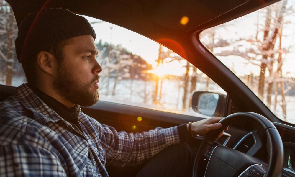 Mann med rutete skjorte og lue holder på rattet og ser på veien mens han kjører på vinterføre. 
