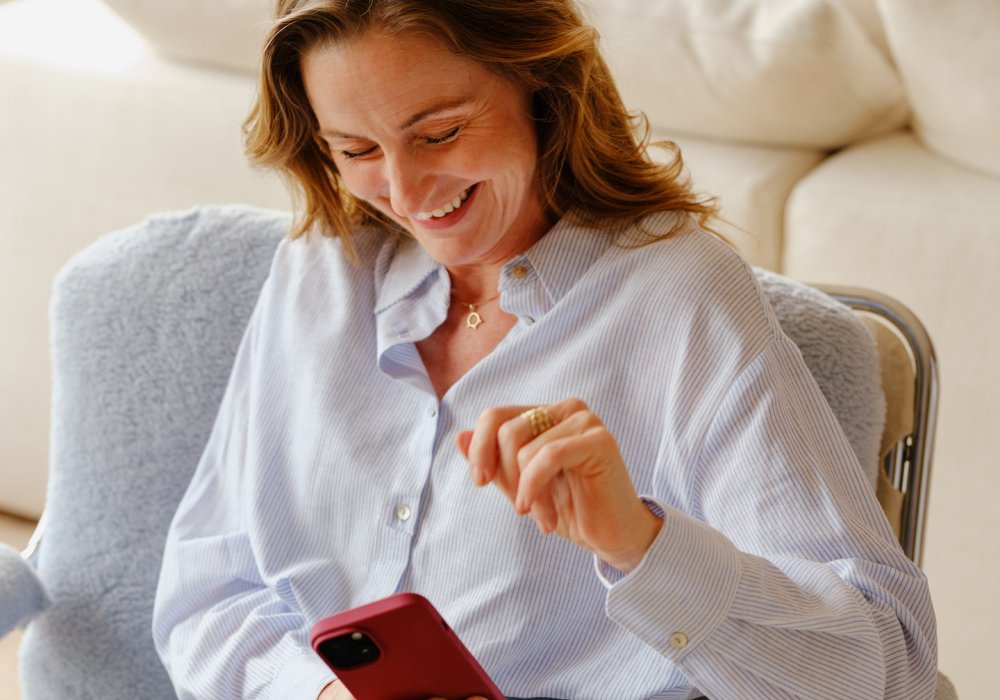 Foto av en smilende dame som sitter i en lenestol og ser på telefonen sin