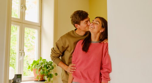 Foto av et ungt par som står i stuen. Mannen kysser sin kjæreste på kinnet Foto av et ungt par som står i stuen. Mannen kysser sin kjæreste på kinnet