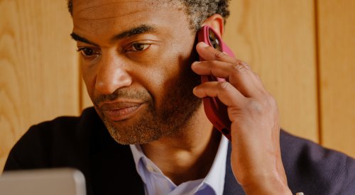 Foto av en mann i dress som sitter å snakker i telefon mens han er på pc-en sin. 