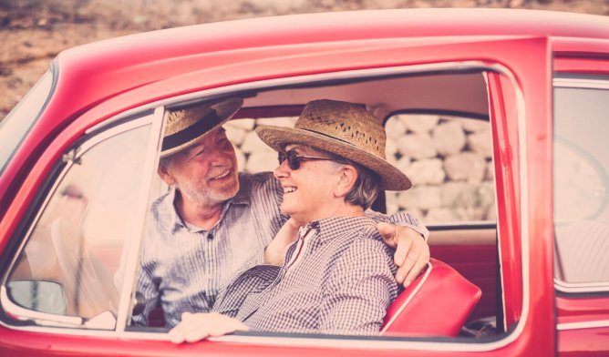 Illustrasjonsfoto. Eldre ektepar som sitter i en rød bil, med hatter. 