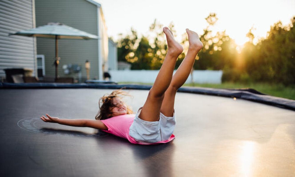 Barn i t-skjorte som lander på rygg på en trampoline. I solnedgang.