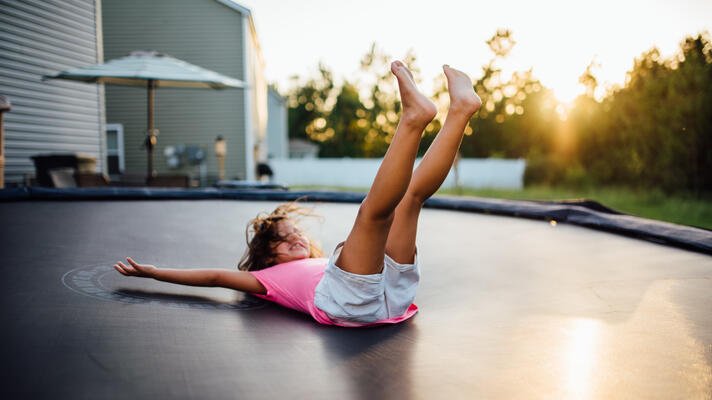 Barn i t-skjorte som lander på rygg på en trampoline. I solnedgang.