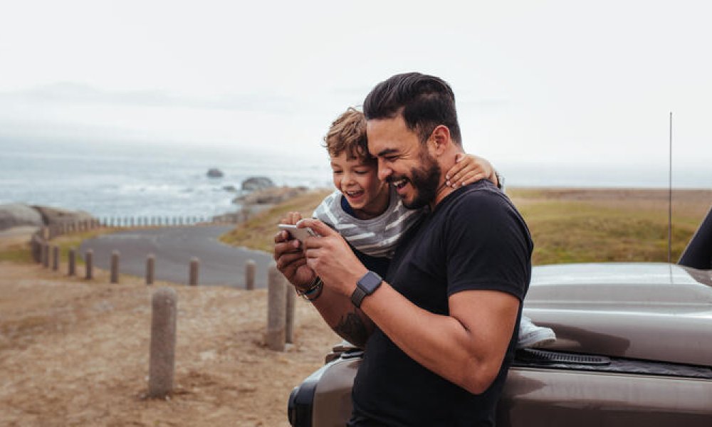 Ung far og hans sønn som titter på en mobil og ler godt. Lener seg på brun bil, ved en strandvei. 