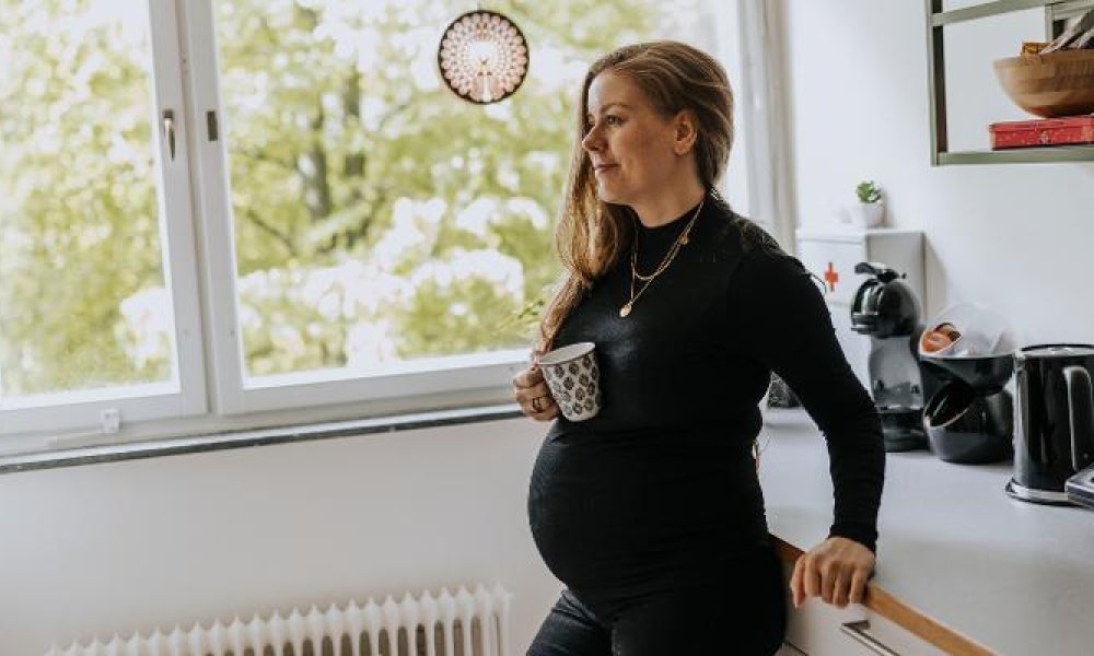 Gravid dame som står på et kjøkken med kaffekopp i hånden.