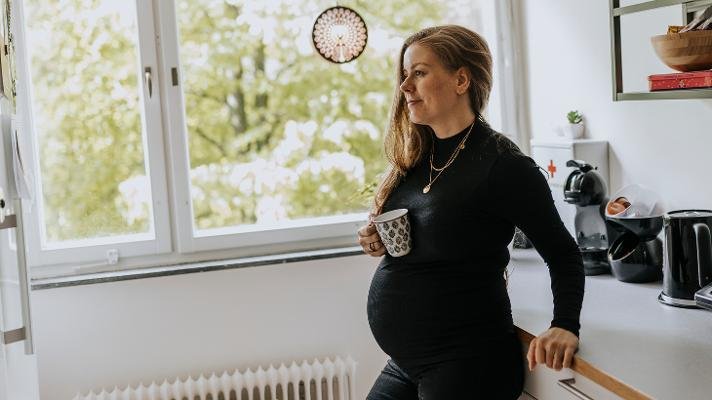 Gravid dame som står på et kjøkken med kaffekopp i hånden.