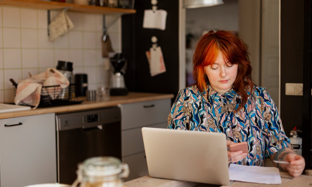 Illustrasjonsfoto: dame med laptop og mobil ved kjøkkenbordet.