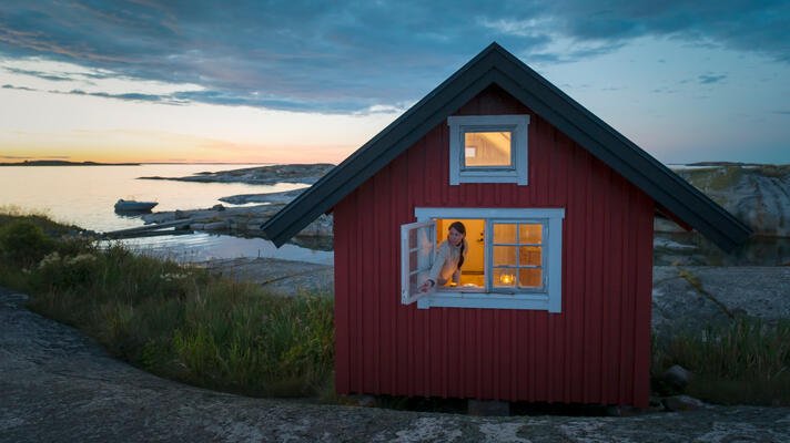 Dame som sitter i vinduet i en rød hytte ved sjøen.