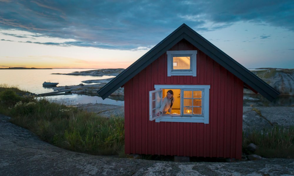 Illustrasjonsfoto: En kvinne åpner vinduet i en liten rød hytte ved sjøen.