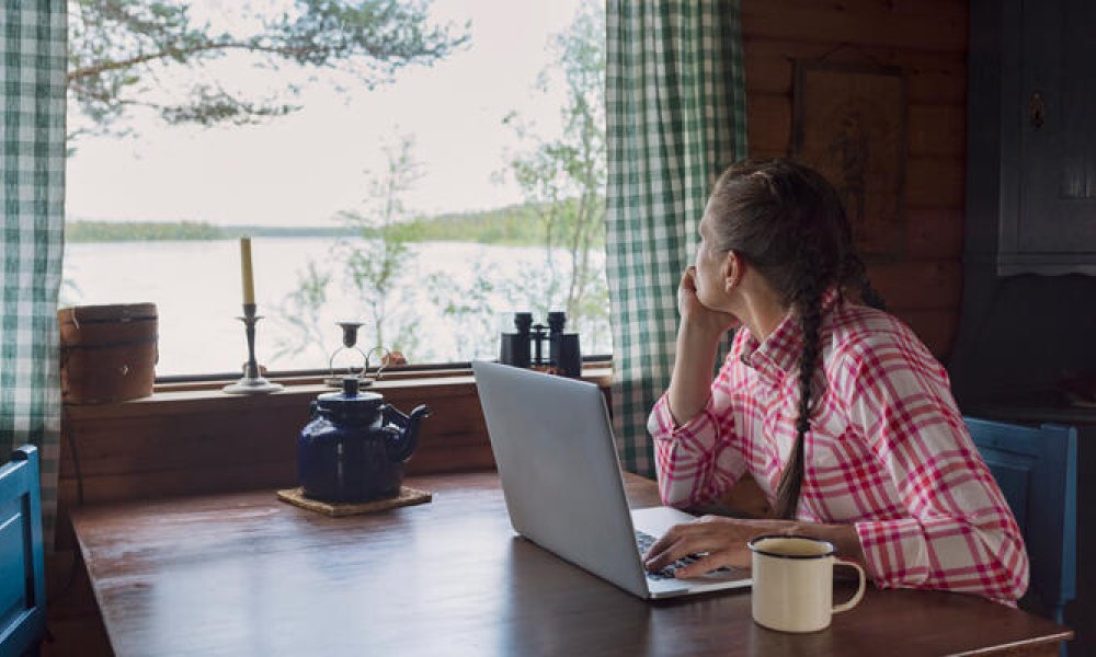 Dame med rødrutete skjorte som sitter ved et spisebord på en hytte og ser ut vinduet.