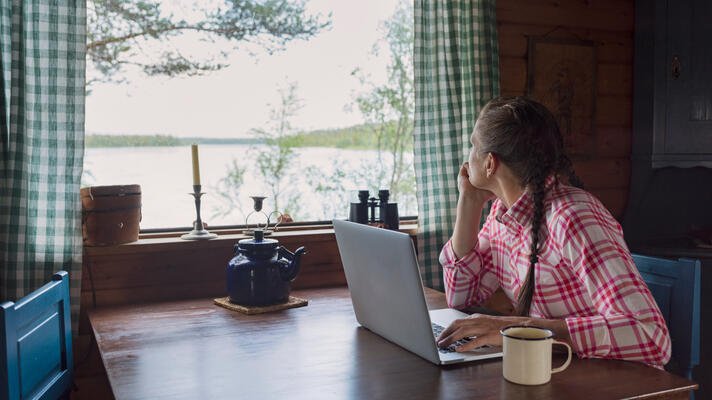 Dame med rødrutete skjorte som sitter ved et spisebord på en hytte og ser ut vinduet.