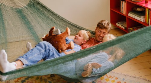 Foto av en far med sine to barn som leker og ler i en hengekøye som henger i stuen Foto av en far med sine to barn som leker og ler i en hengekøye som henger i stuen