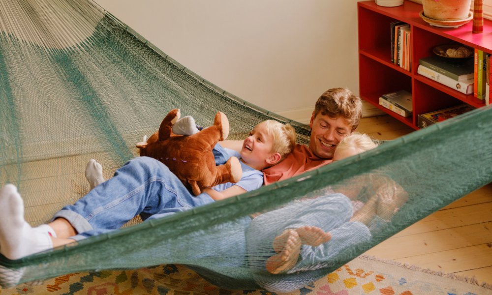 Foto av en far med sine to barn som leker og ler i en hengekøye som henger i stuen