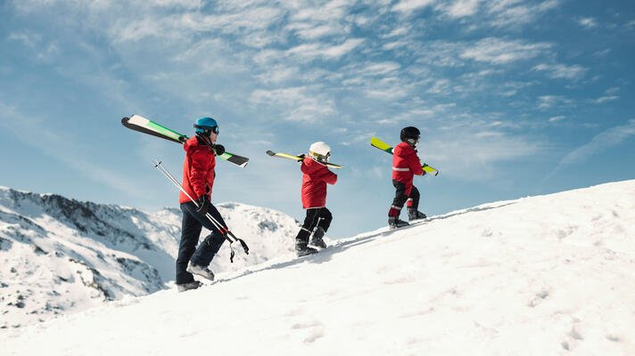 Tre familiemedlemmer som går opp en fjellrygg av snø. Ski på skuldrene. 