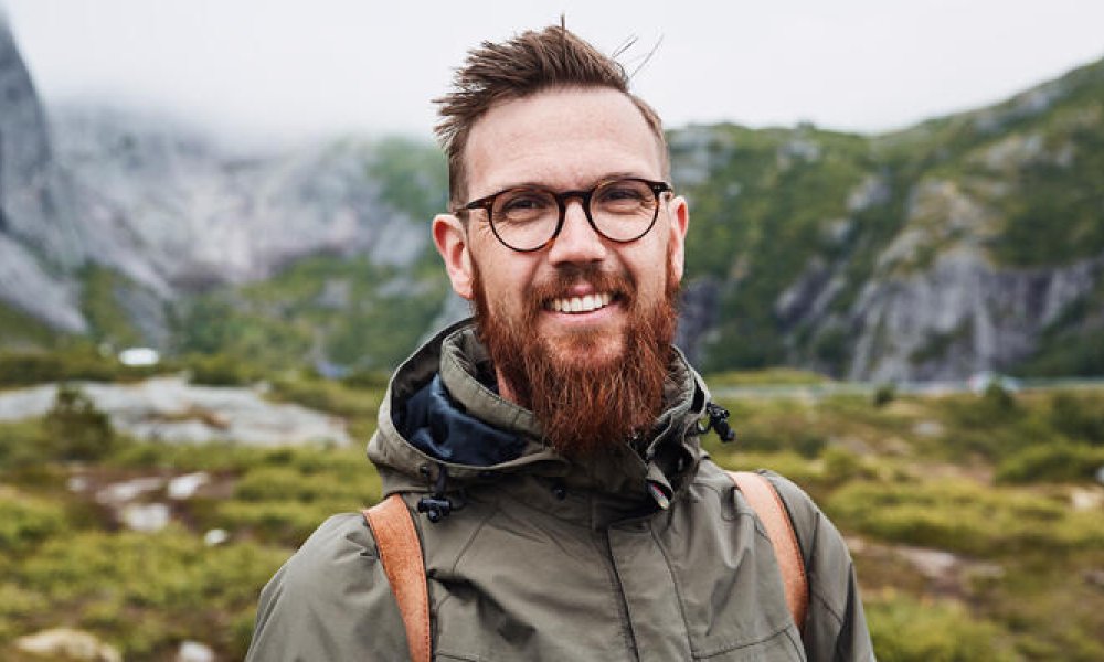 Portrettfoto av ung mann på tur i naturen med rødt skjegg, briller og skinnryggsekk.