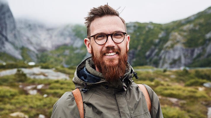 Portrettfoto av ung mann på tur i naturen med rødt skjegg, briller og skinnryggsekk.