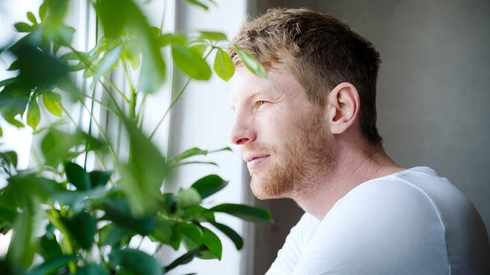 Mann i hvit t-skjorte titter ut av vindu med plante i forgrunnen.