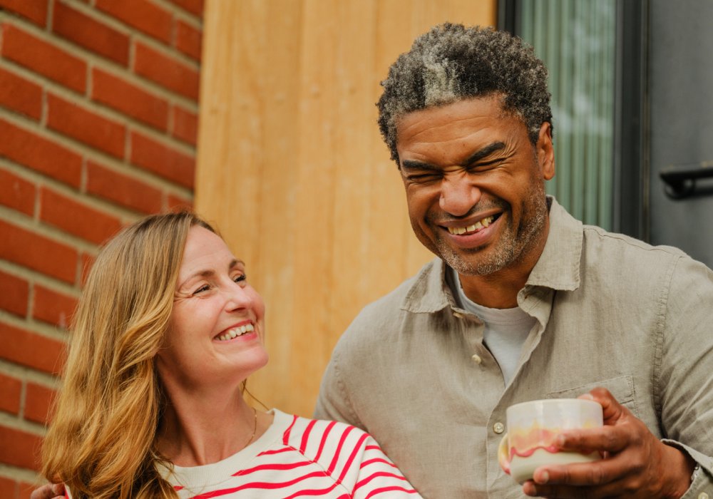 Foto av et smilende par som sitter i trappeoppgangen til et hus og holder hver sin kaffekopp Foto av et smilende par som sitter i trappeoppgangen til et hus og holder hver sin kaffekopp