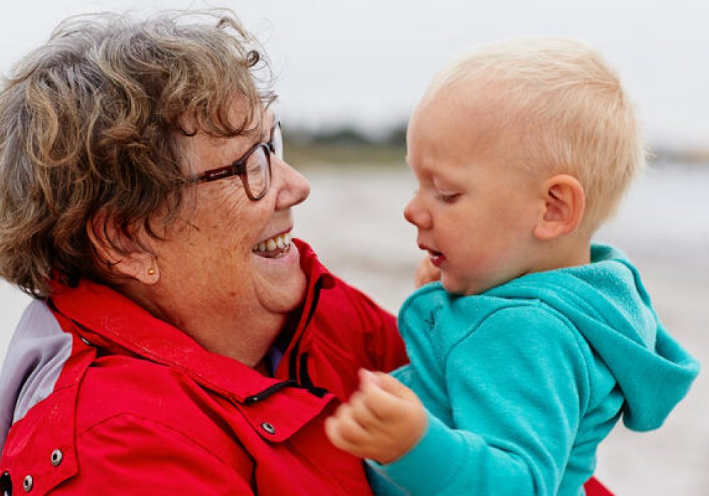 Bestemor i rød jakke holder baby på strand.