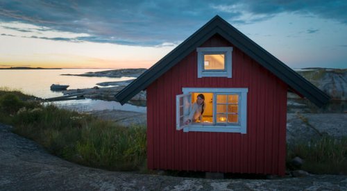 Dame som sitter i vinduet i en rød hytte ved sjøen.