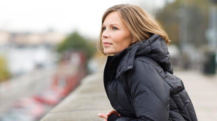 Dame i sort boblejakke som står på en bro og ser utover.