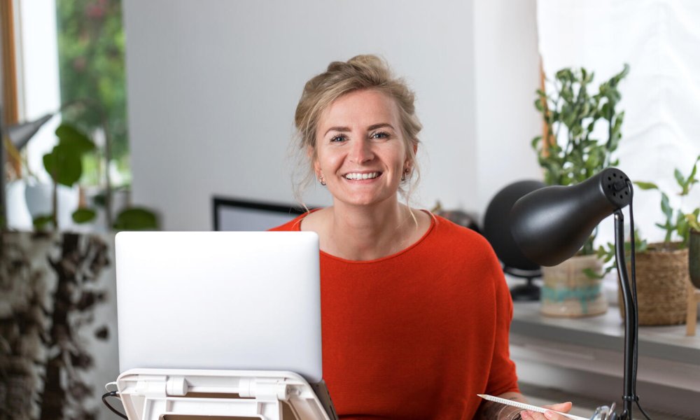 Illustrasjonsfoto: Dame i rød genser sitter foran PC på kontor mens hun smiler til kamera