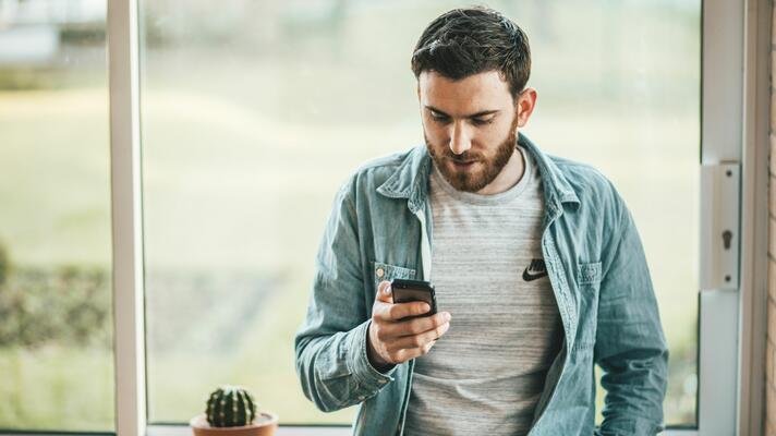 Mann i blå dongeriskjorte som står ved et vindu og sjekker mobilen sin.